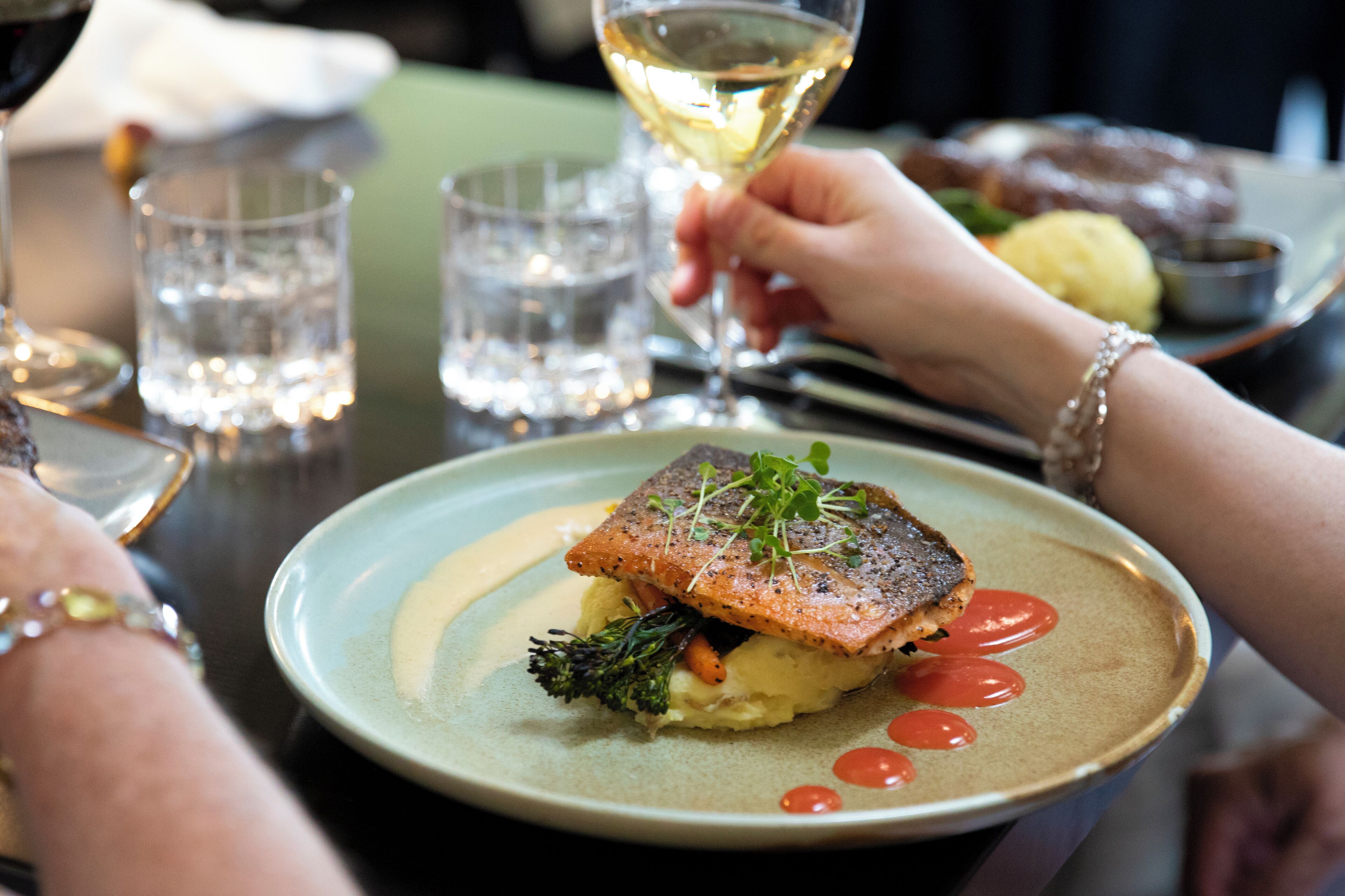 Une table à manger avec une assiette de nourriture, des verres d’eau et une personne tenant un verre de vin blanc.