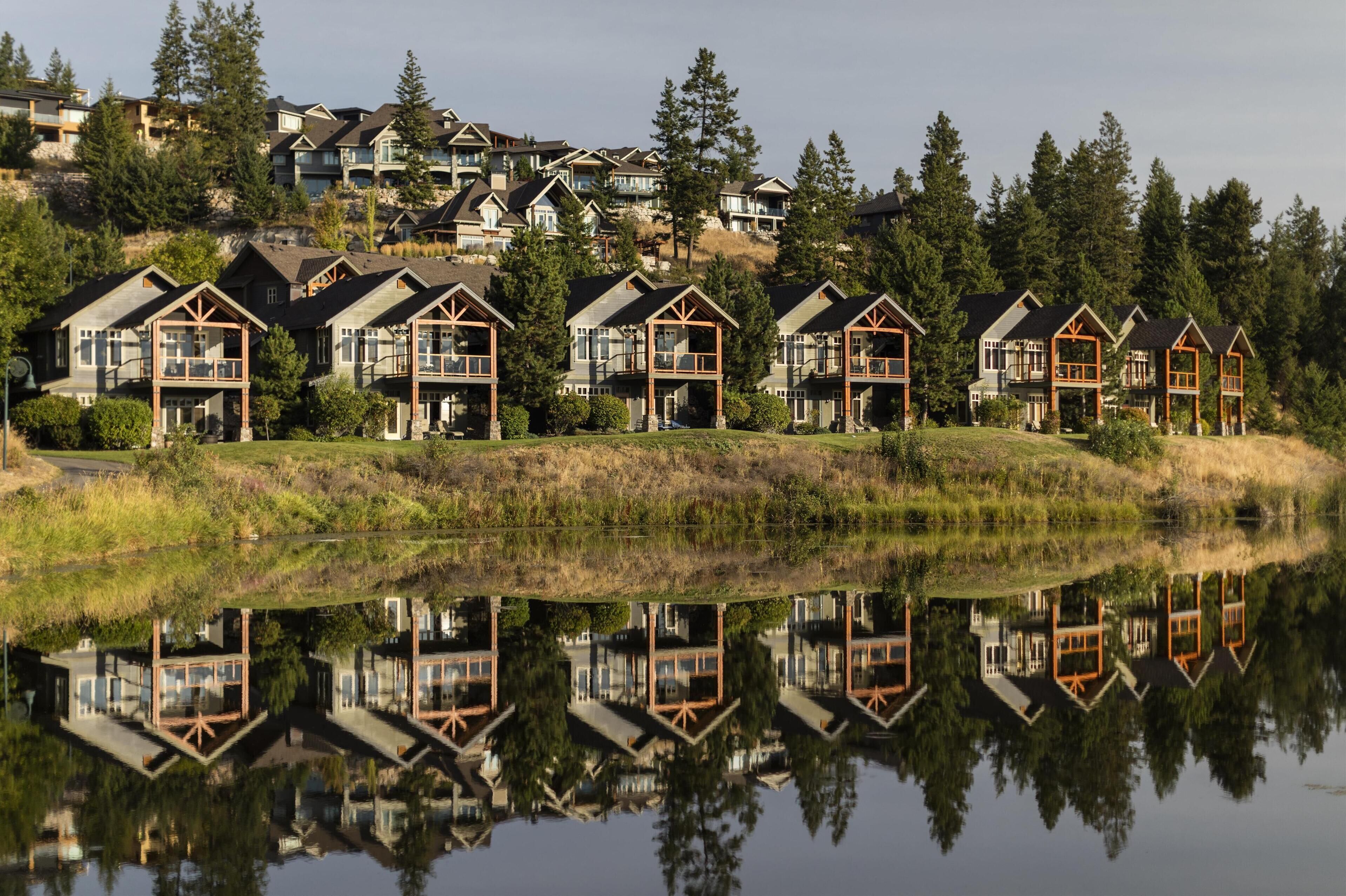 A row of modern houses with large windows and wooden balconies, reflecting in a calm body of water.
