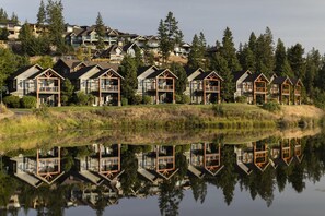 A row of modern houses with large windows and wooden balconies, reflecting in a calm body of water.