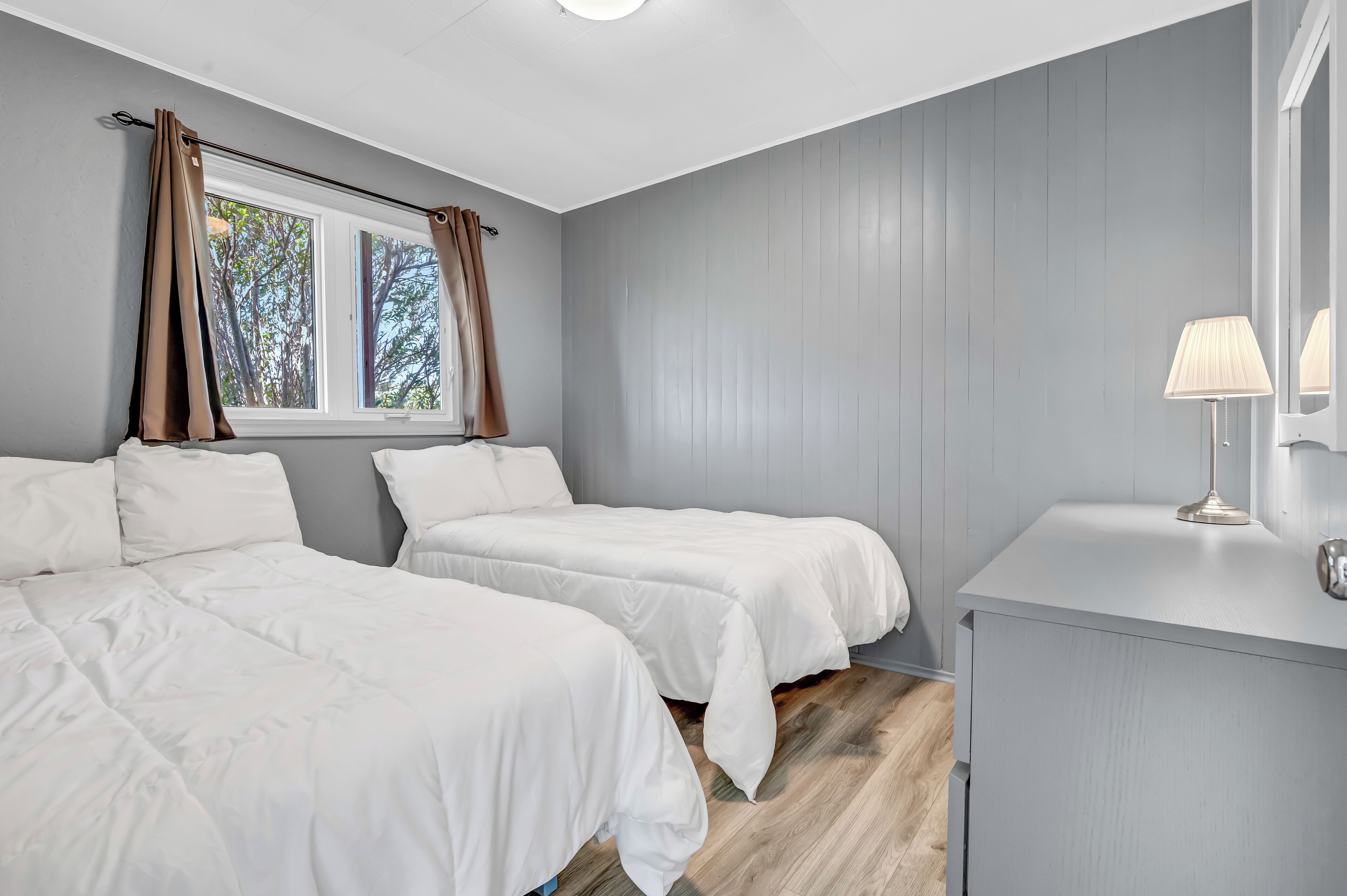 Quarto com duas camas, uma janela com cortinas, mesa de cabeceira com abajur e piso de madeira.