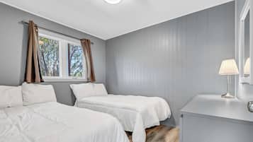 A bedroom with two beds, a window with curtains, a bedside table with a lamp, and a wooden floor.