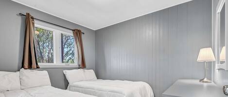 Quarto com duas camas, uma janela com cortinas, mesa de cabeceira com abajur e piso de madeira.