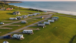A campground with RVs parked on green fields, a beach, and houses in the background. - Mavillette Beach Lodge and RV Park (Mavillette)