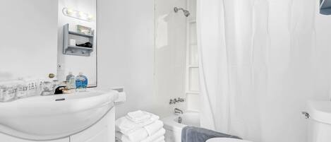 Un baño moderno con lavamanos, espejo y tocador blancos, una ducha con cortina y un inodoro.