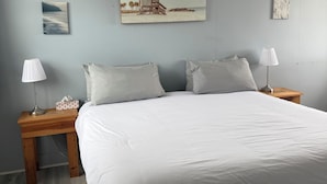 A bedroom with a large bed, two bedside tables with lamps, a wooden headboard, and a beach scene painting on the wall. - Mavillette Beach Lodge and RV Park (Mavillette)