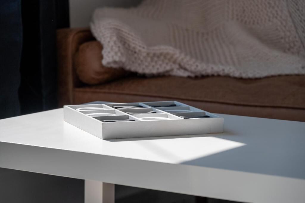 Une table blanche sur laquelle se trouve un objet blanc, un canapé marron avec une couverture blanche en arrière-plan, et une fenêtre qui illumine la scène.