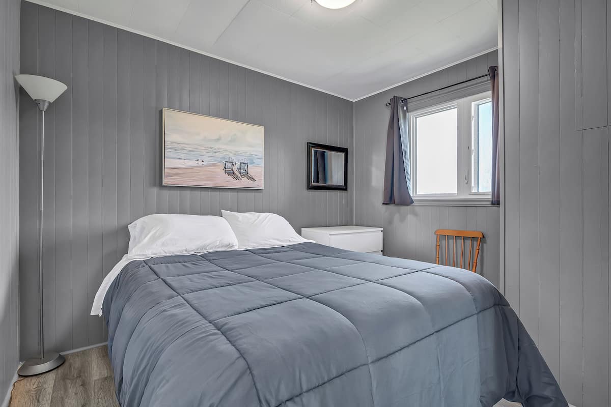 Ein Schlafzimmer mit einem Bett, einer Nachttischlampe, einem Fenster mit Vorhängen, einem gerahmten Bild und einem kleinen Holzstuhl.
