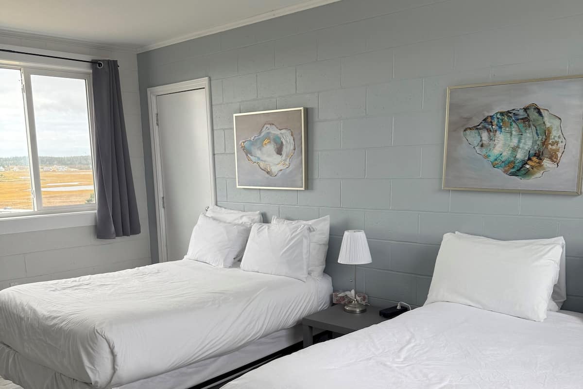 Ein Hotelzimmer mit zwei Betten, einem Fenster mit Vorhängen, einer Tür und zwei an der Wand befestigten Kunstwerken.