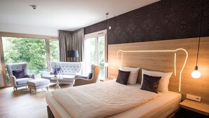 A modern hotel room with a large bed, a sofa, two armchairs, a small table, and a large window offering a view of greenery. - Parkhotel Schillerhain (Kirchheimbolanden)