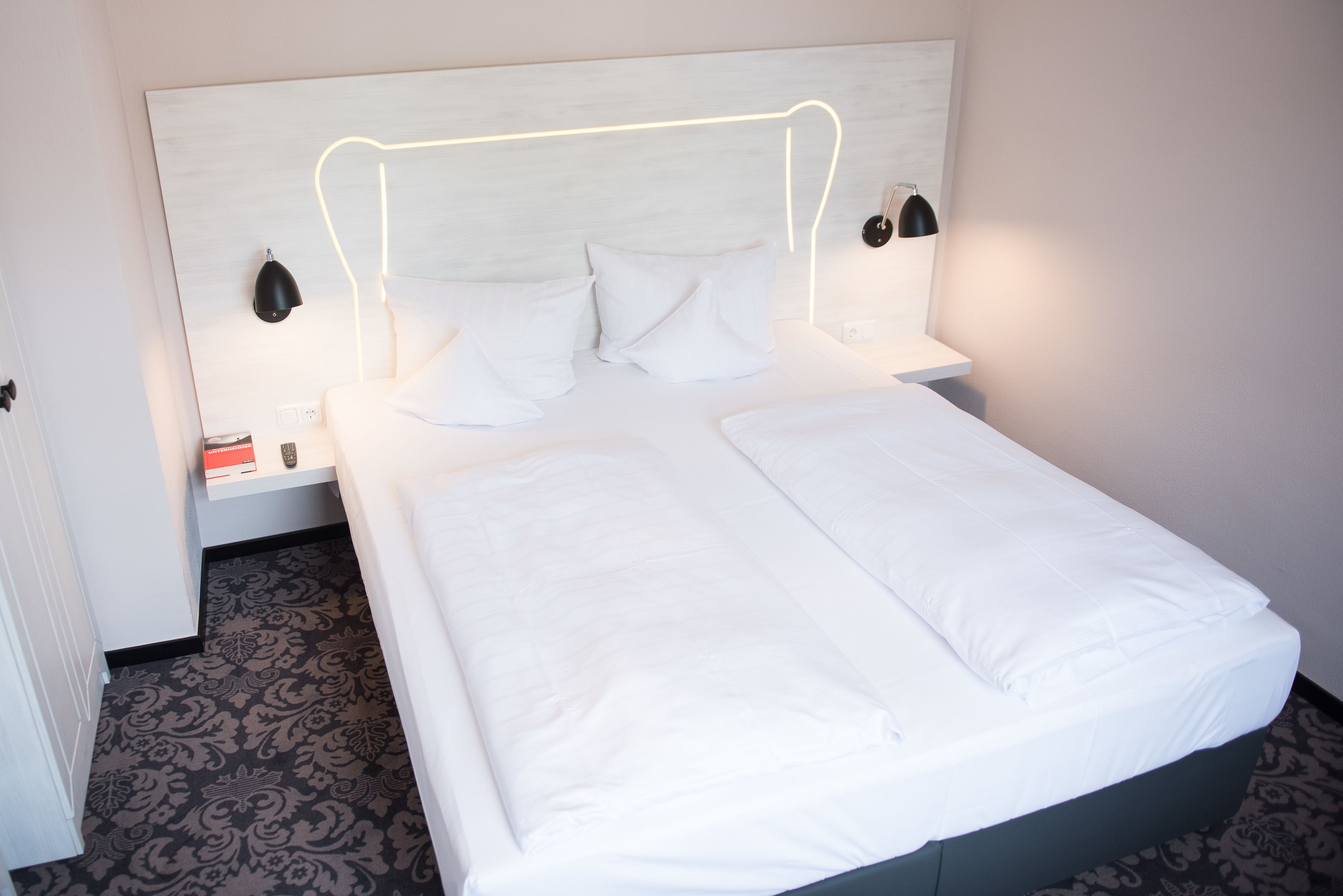Un lit bien fait, avec du linge de lit blanc, une tête de lit dotée d’un éclairage intégré et des lampes de chevet.
