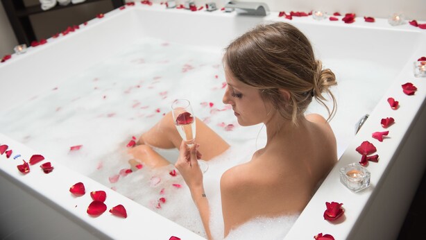 A woman in a bathtub filled with bubbles and rose petals, holding a glass of wine. - Parkhotel Schillerhain (Kirchheimbolanden)