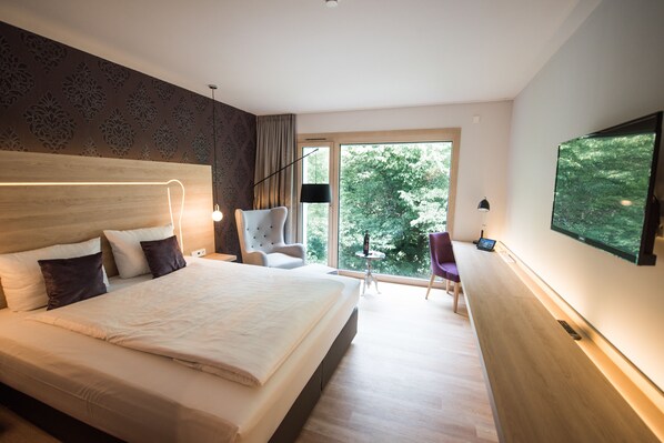 A modern hotel room with a large bed, a desk, a chair, and a large window offering a view of trees. - Parkhotel Schillerhain (Kirchheimbolanden)