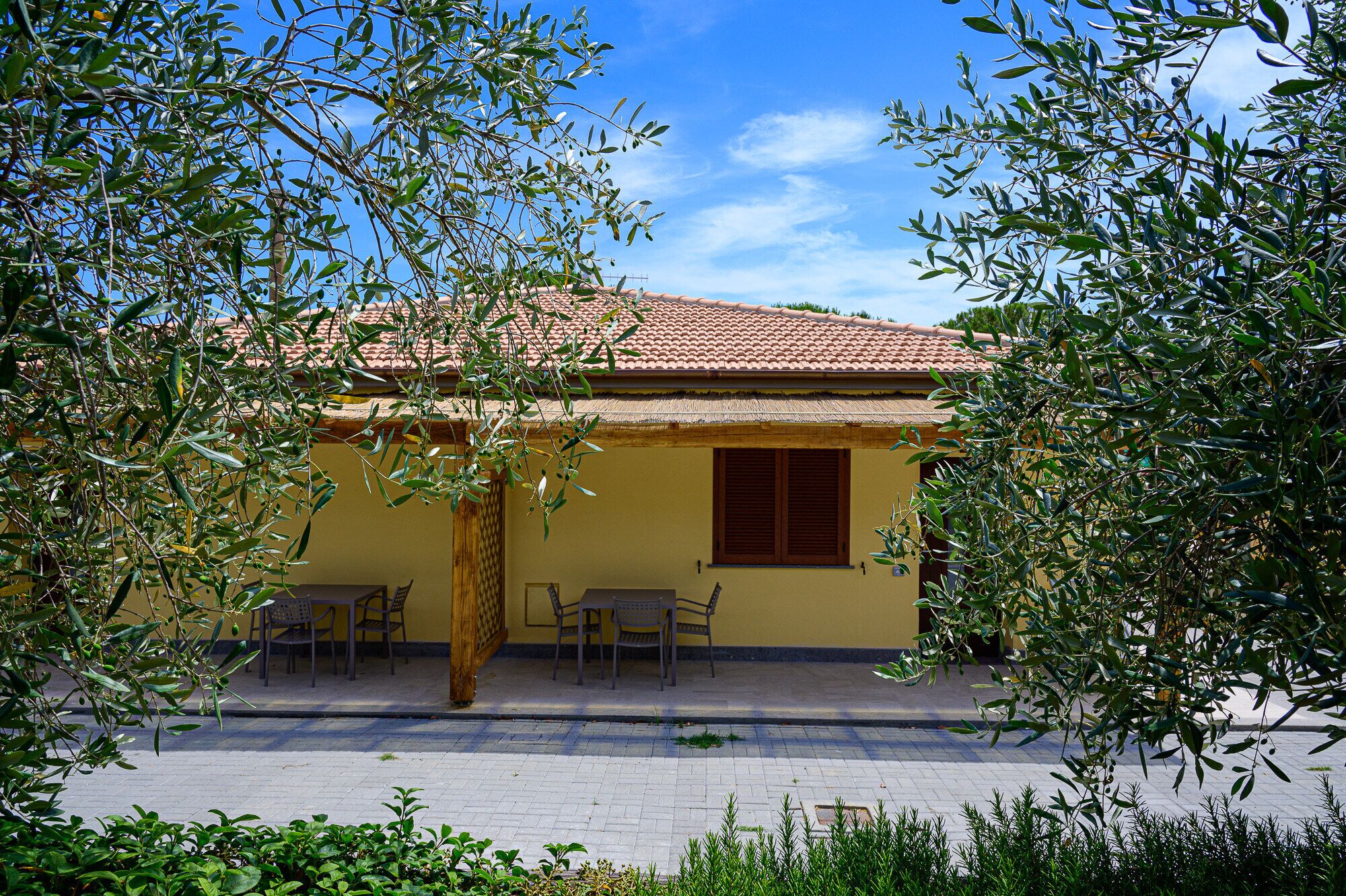 A yellow building with a tiled roof, a window with wooden shutters, and a patio with chairs.