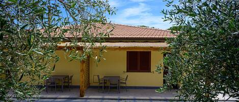 A yellow building with a tiled roof, a window with wooden shutters, and a patio with chairs.