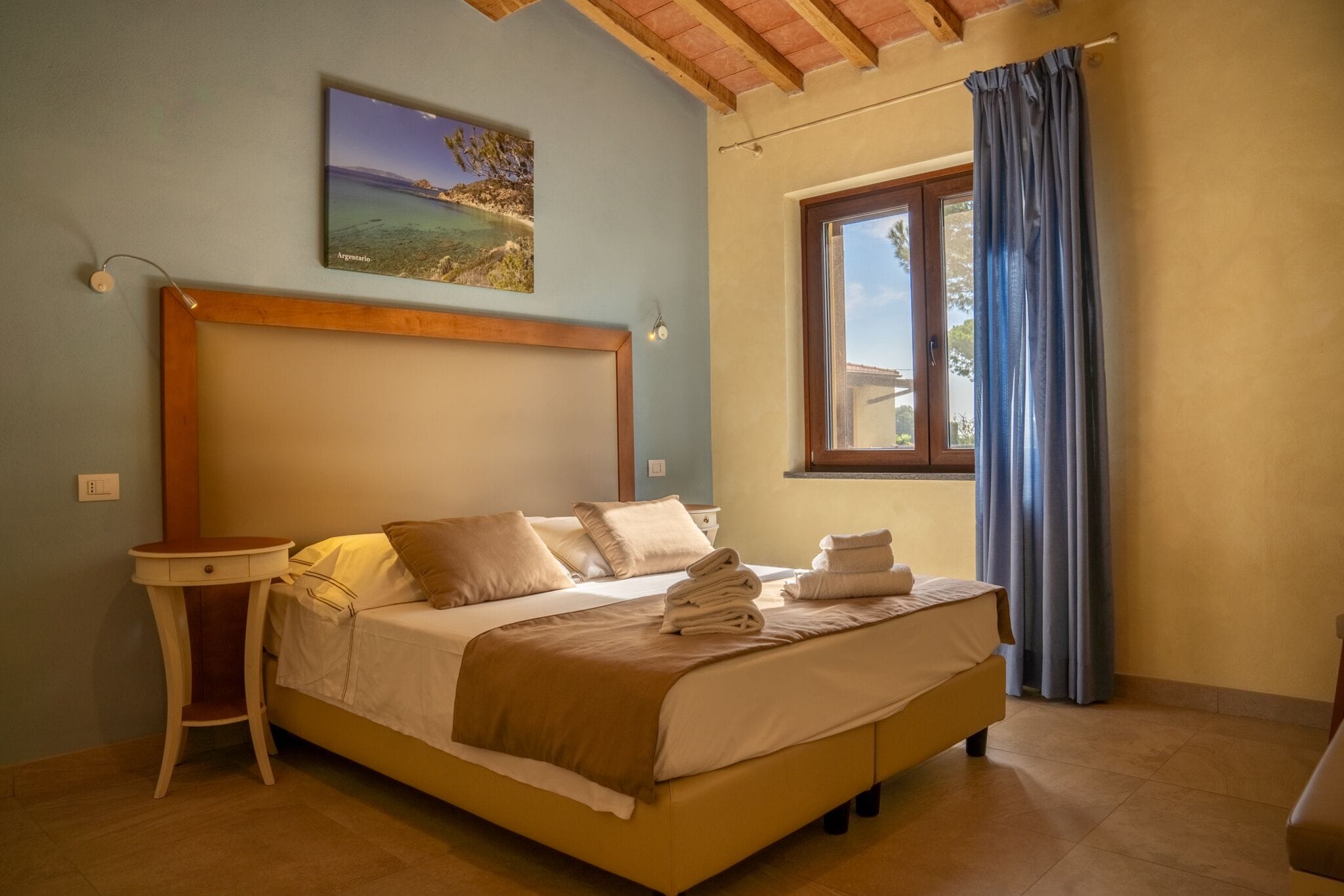 A bedroom with a bed, a bedside table, a window with curtains, and a painting on the wall.
