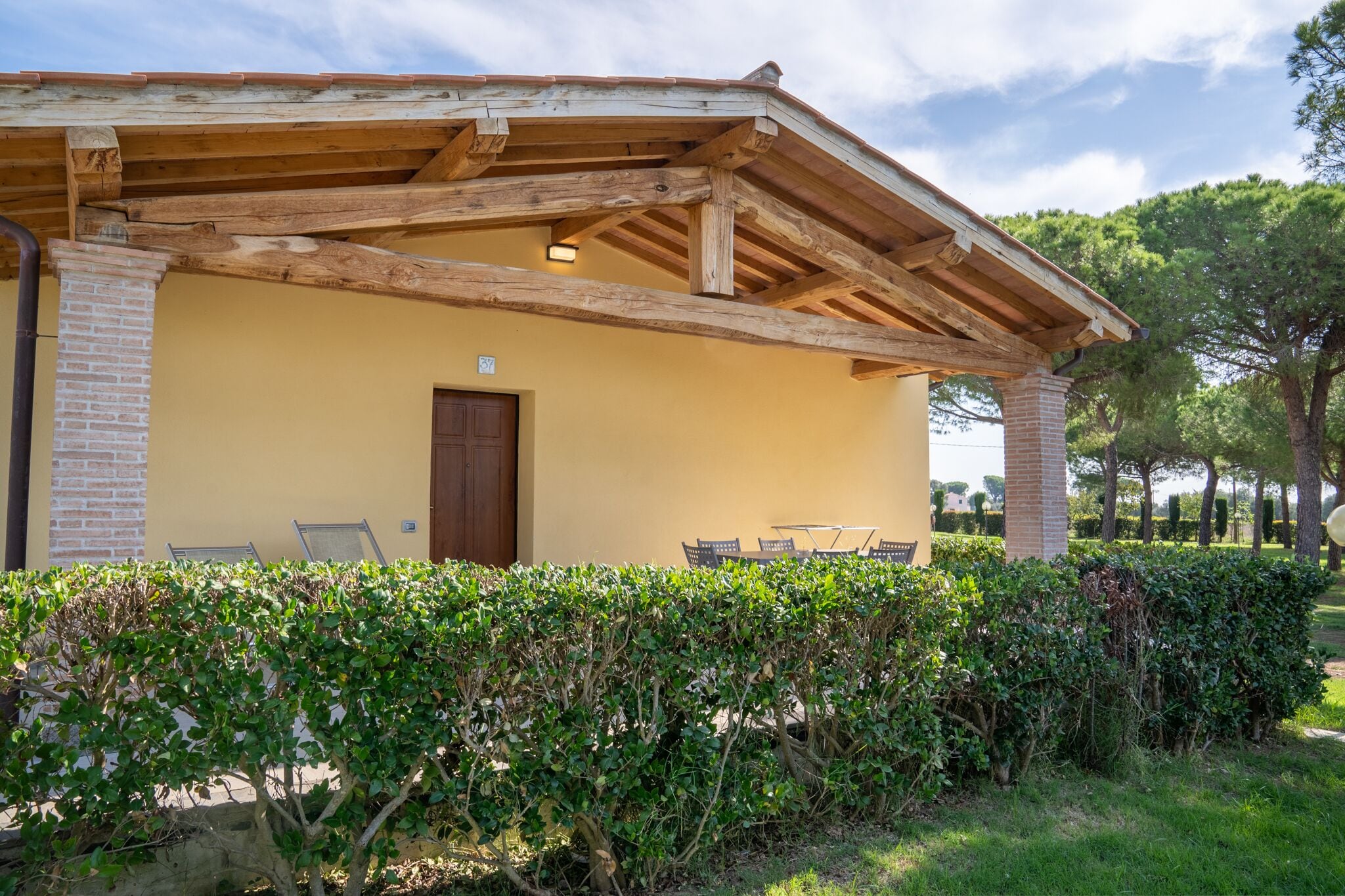 Un edificio con una pérgola de madera, una chimenea de ladrillo y una puerta. Hay sillas y una mesa al aire libre.