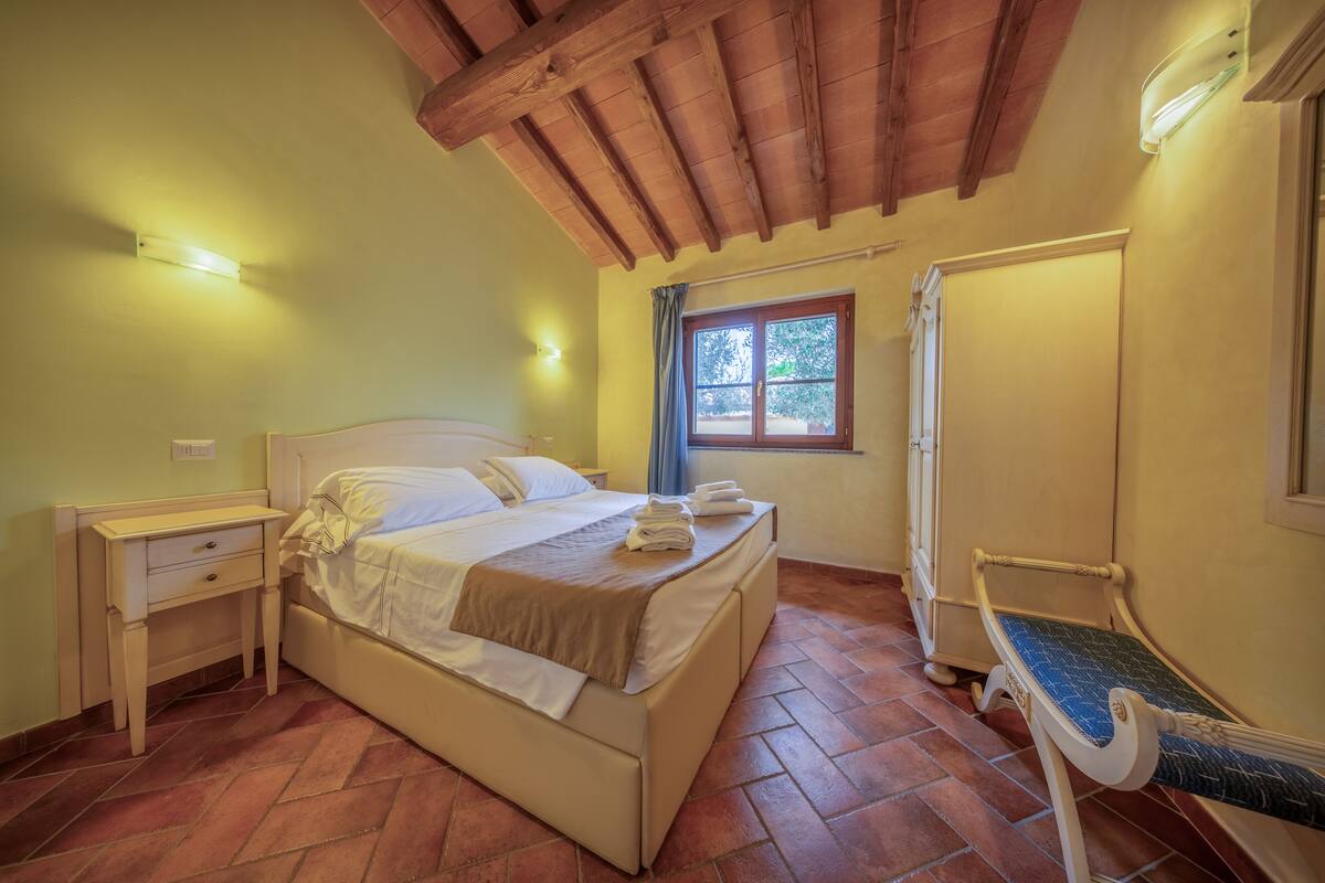 A bedroom with a bed, a nightstand, a bench, a window, and a door.