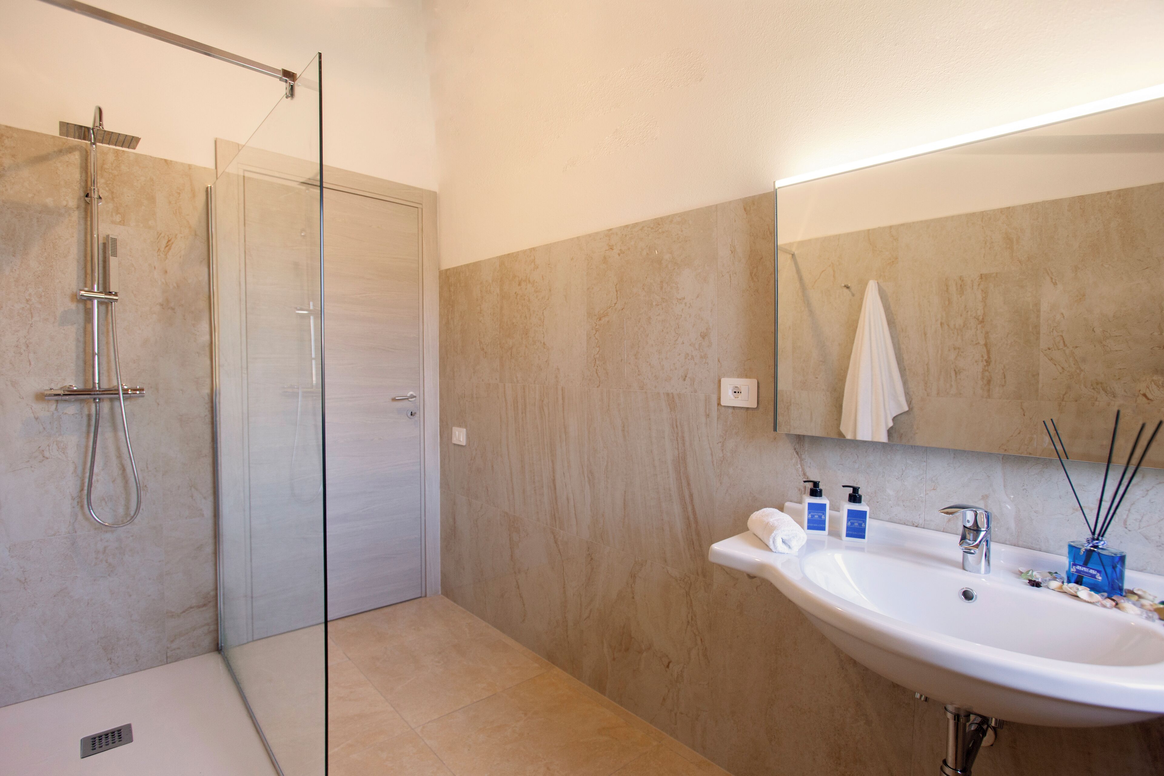 A modern bathroom with a glass-enclosed shower, a large sink with a mirror, and beige marble walls.