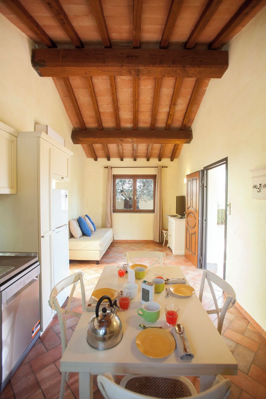 Un comedor con una mesa lista para dos, una cocina con refrigerador y armarios, un sofá con cojines, y una ventana con cortinas.