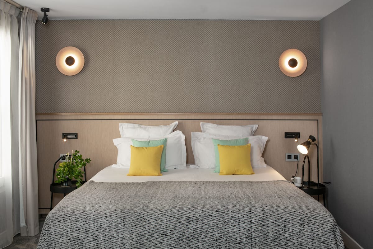 Quarto de hotel com uma cama grande, duas mesas de cabeceira, um criado-mudo, uma luminária e um vaso com planta.