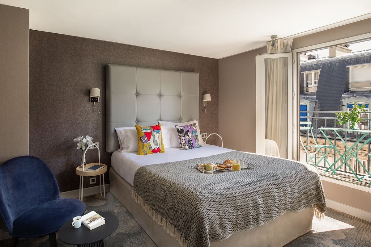 Quarto de hotel com uma cama grande, uma cadeira azul, uma mesa pequena com uma xícara e um livro, e uma varanda com corrimão e uma planta.