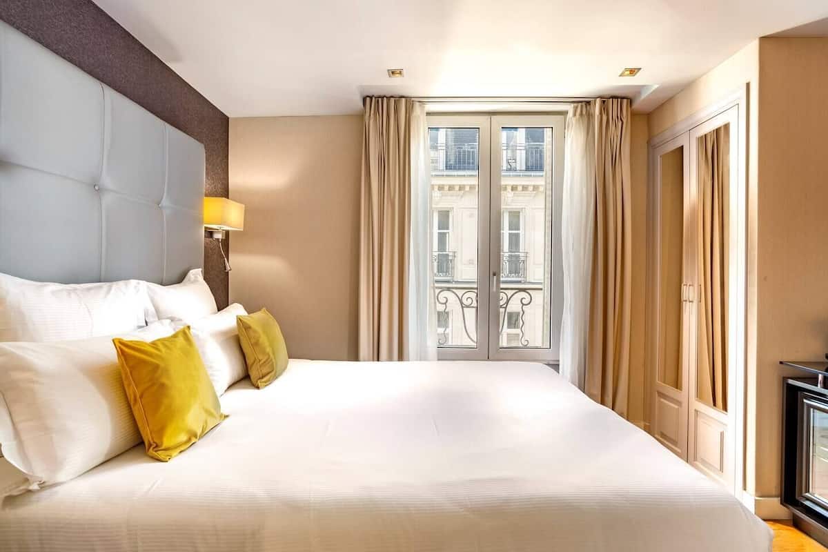 Quarto de hotel com uma cama grande, cabeceira, mesa de cabeceira, abajur, janela com cortinas e vista para edifícios.
