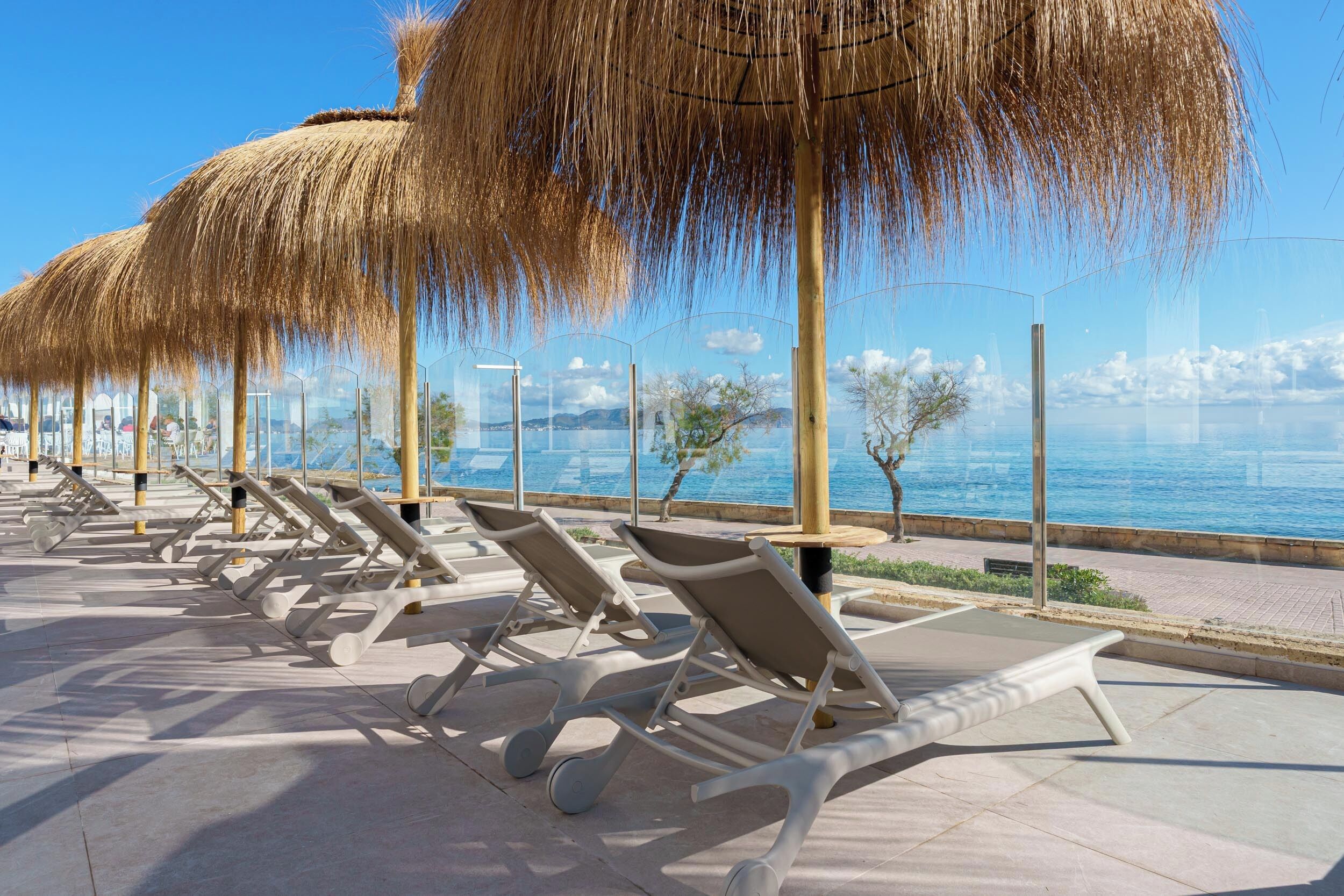 A row of sun loungers with thatched umbrellas, a clear view of the sea, and a paved walkway.