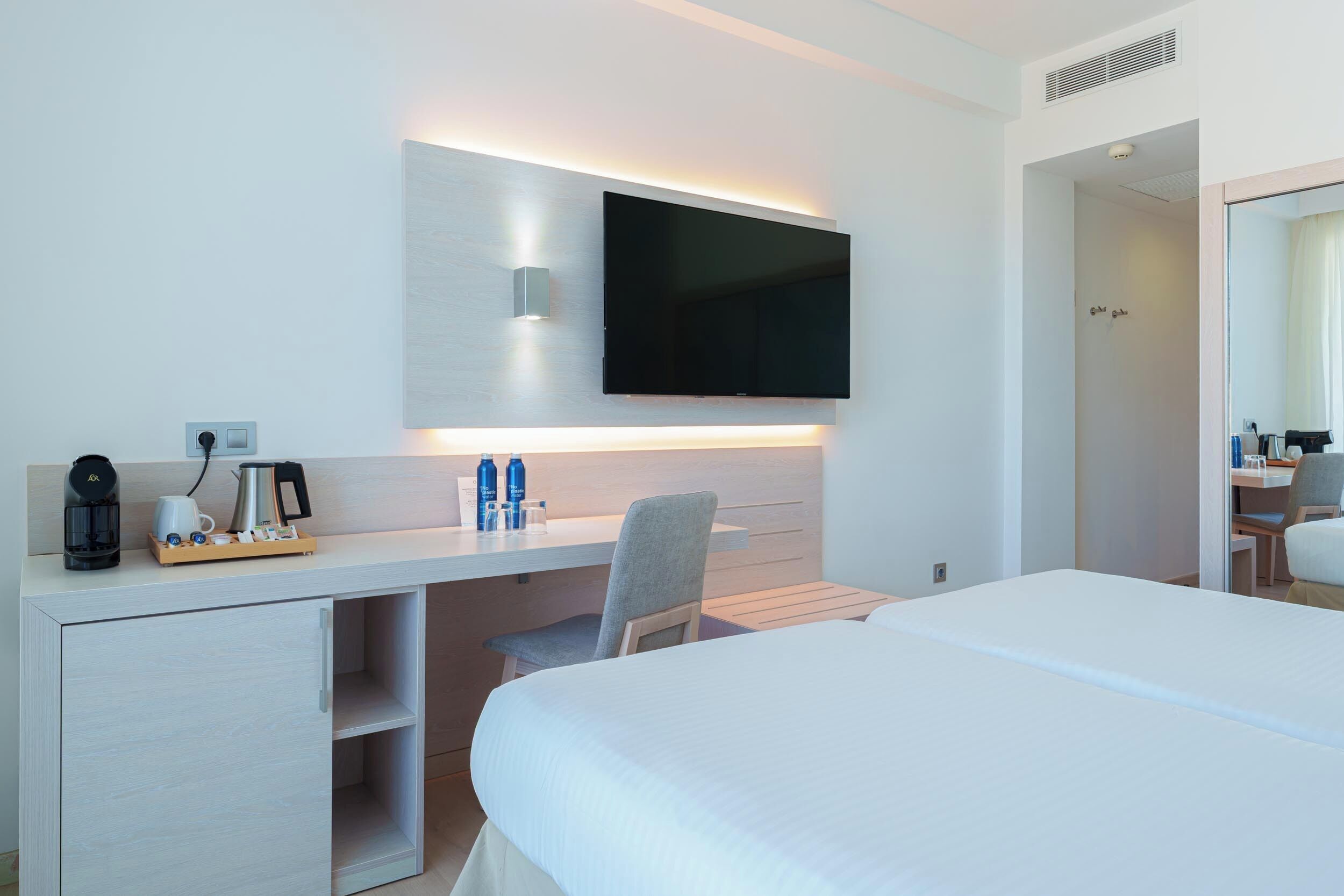 A modern hotel room with a bed, a desk with a flat-screen TV, a coffee machine, and a mirror.