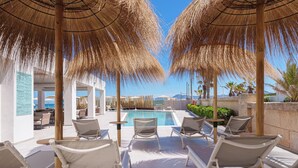 A poolside area with lounge chairs and umbrellas, a pool, and a view of the sea and mountains.