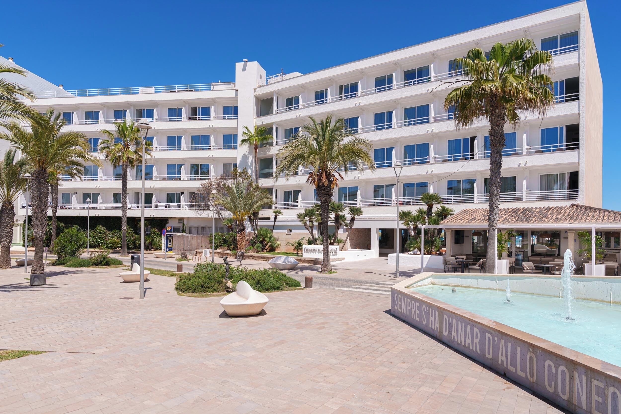 Ein mehrstöckiges Hotel mit Poolbereich, Palmen und Außenmöbeln.