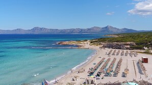 A beach with rows of umbrellas and lounge chairs, a rocky shoreline, and mountains in the background.
