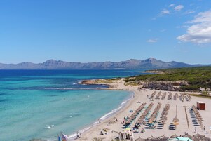 Una playa con hileras de sombrillas y sillas de descanso, un litoral rocoso y montañas al fondo.