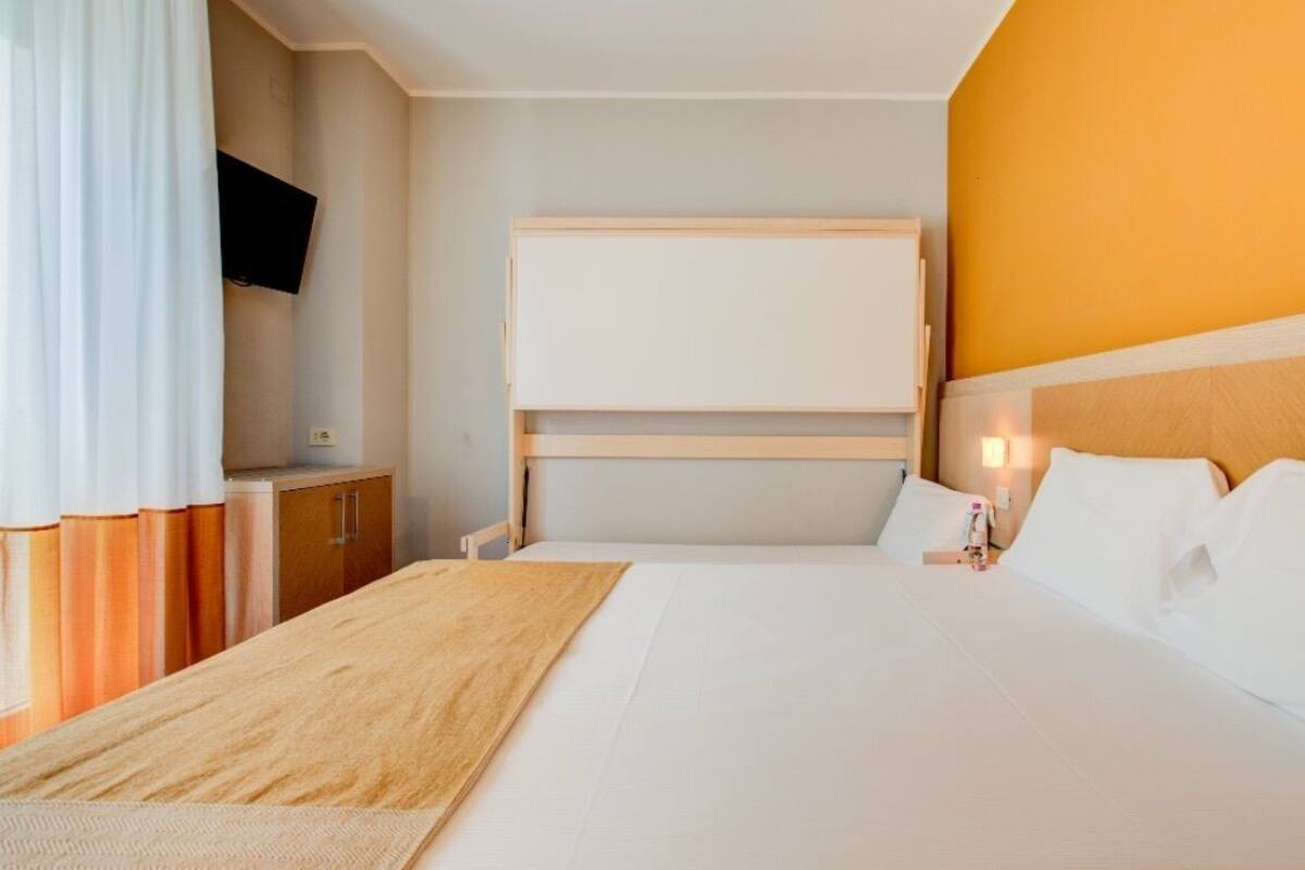 A hotel room with a bed, a flat-screen TV mounted on the wall, a wooden headboard, and a bedside table with a lamp.