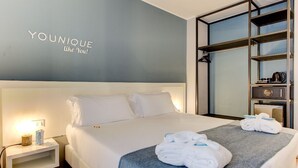 A hotel room with a bed, a nightstand, a wall-mounted shelf with a coffee maker, and a towel on the bed. - Polo BeYou Hotel (Riccione)