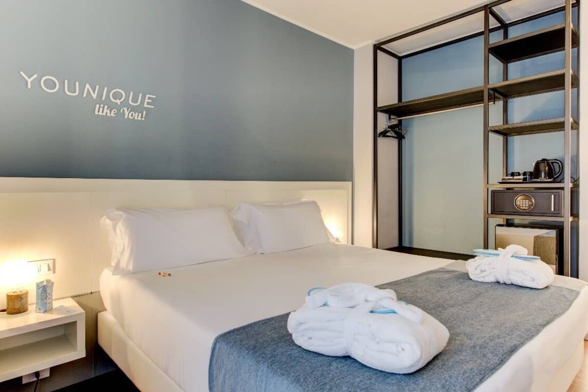 A hotel room with a bed, a nightstand, a wall-mounted shelf with a coffee maker, and a towel on the bed.