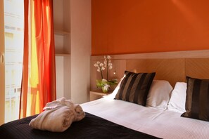 Una habitación de hotel con cama, almohadas, una mesita de noche, un florero con flores y una ventana con cortinas.