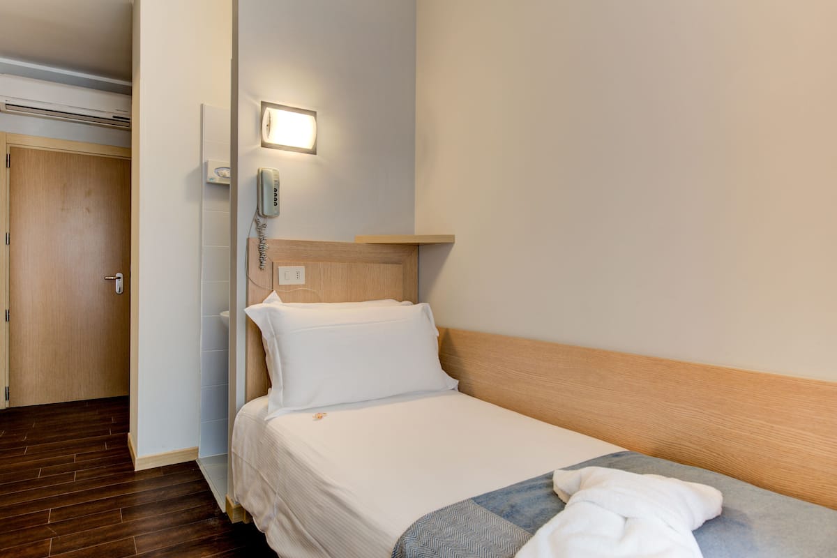 A hotel room with a wooden headboard, a single bed with white bedding, a bedside table, a wall-mounted phone, and a door to the left.