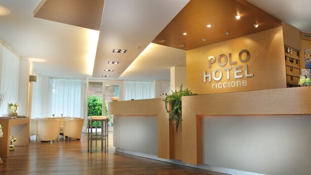 A hotel reception area with a wooden reception desk, a seating area with chairs, and a view of the outdoors. - Polo BeYou Hotel (Riccione)