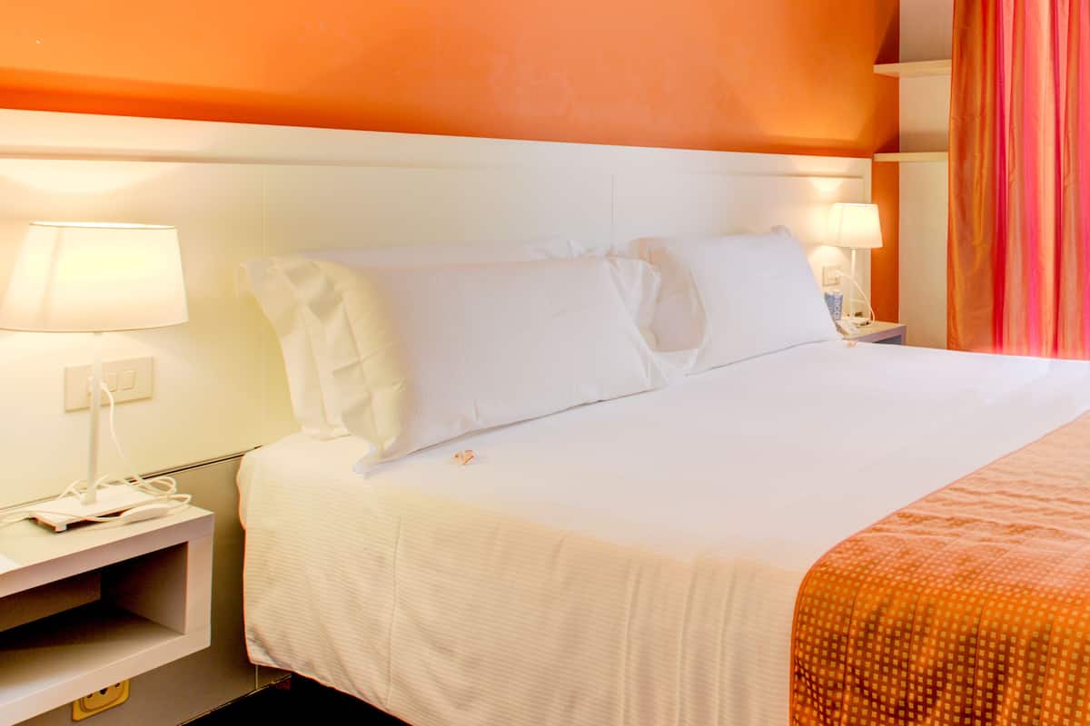 A hotel room with a large bed, white bedding, a nightstand with a lamp, and an orange accent wall.