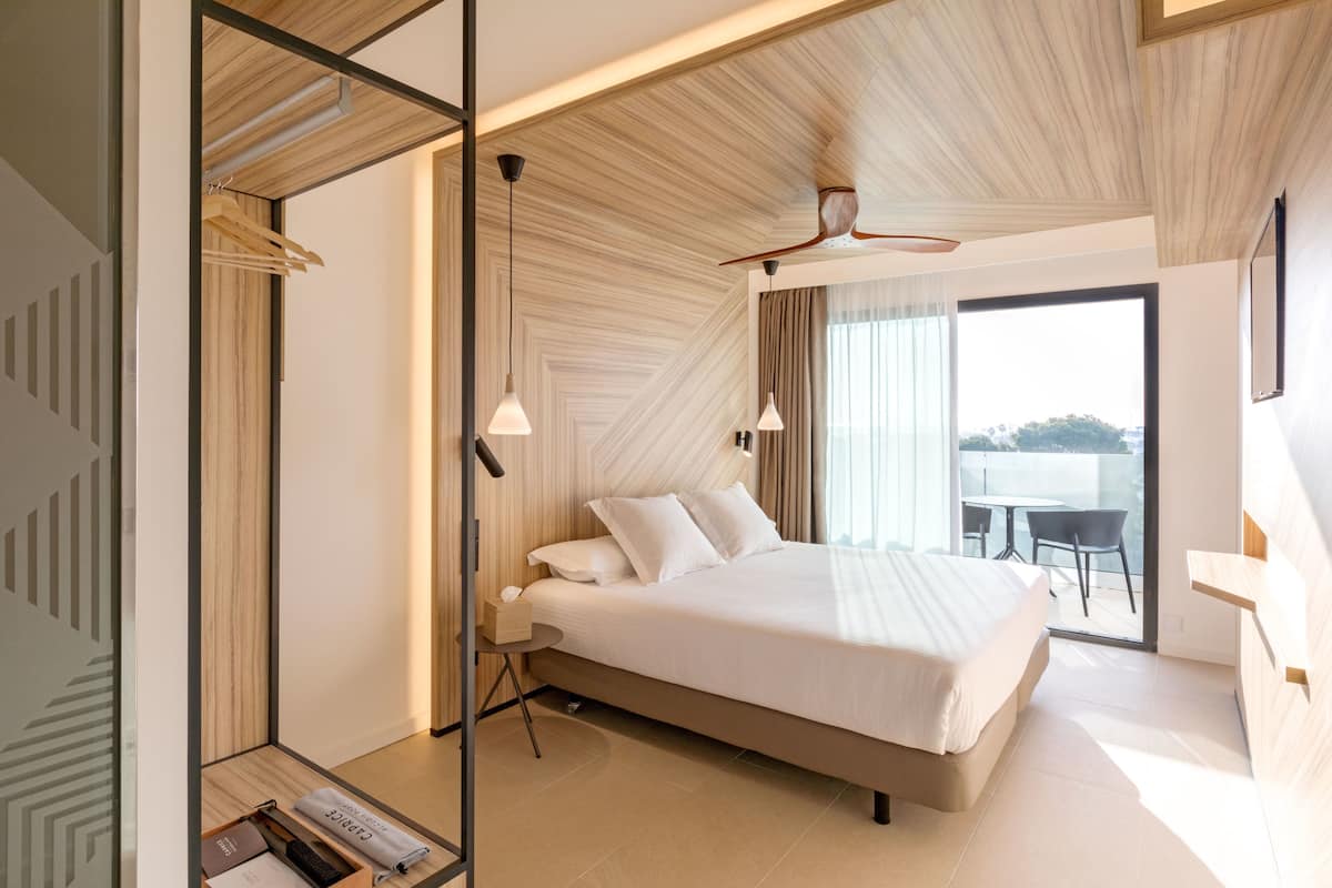 A modern hotel room with a large bed, a small side table, a ceiling fan, and a view of the outdoors through a large window.