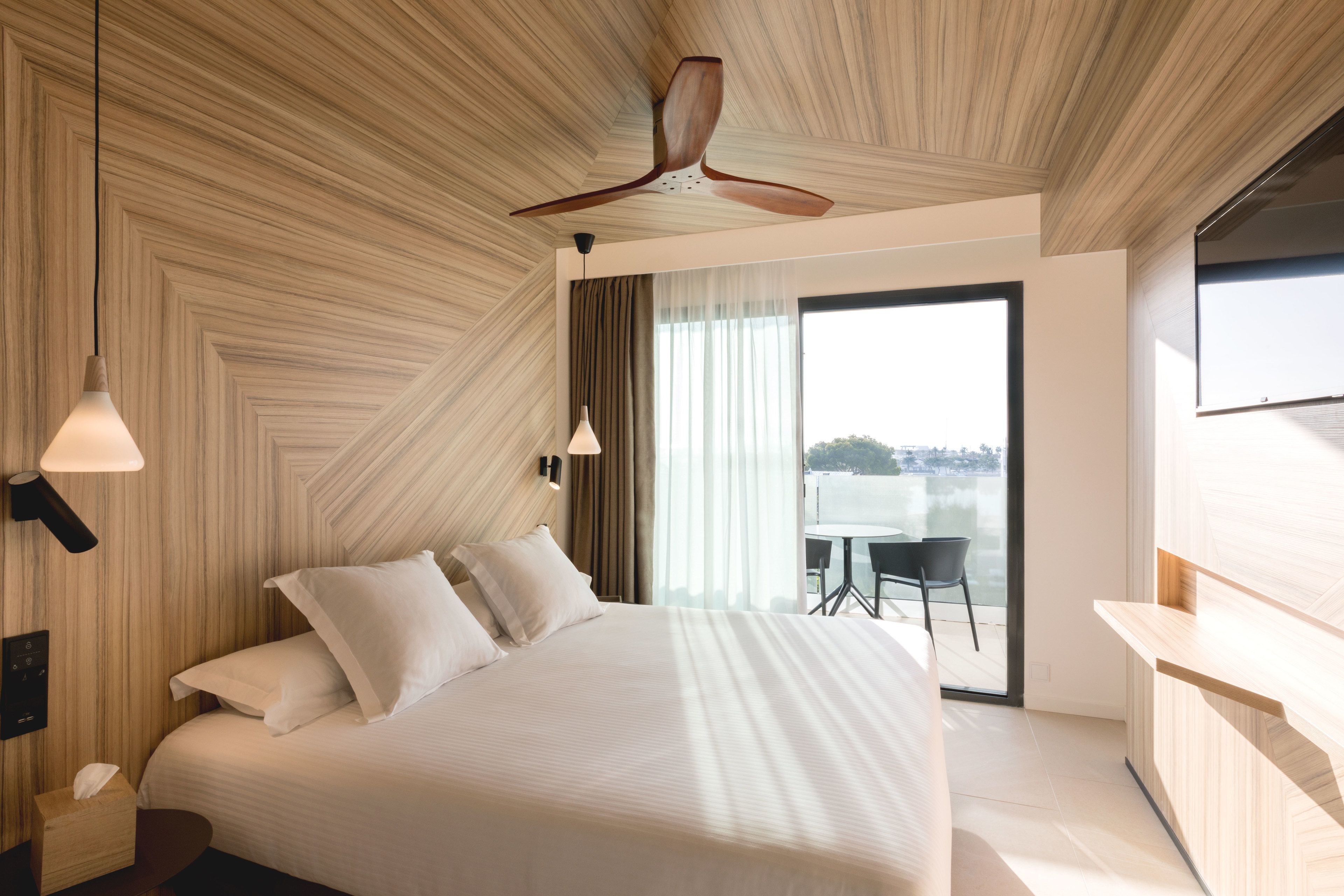 Una habitación de hotel moderna con una cama grande, un ventilador de techo y vistas a la ciudad a través de amplios ventanales.