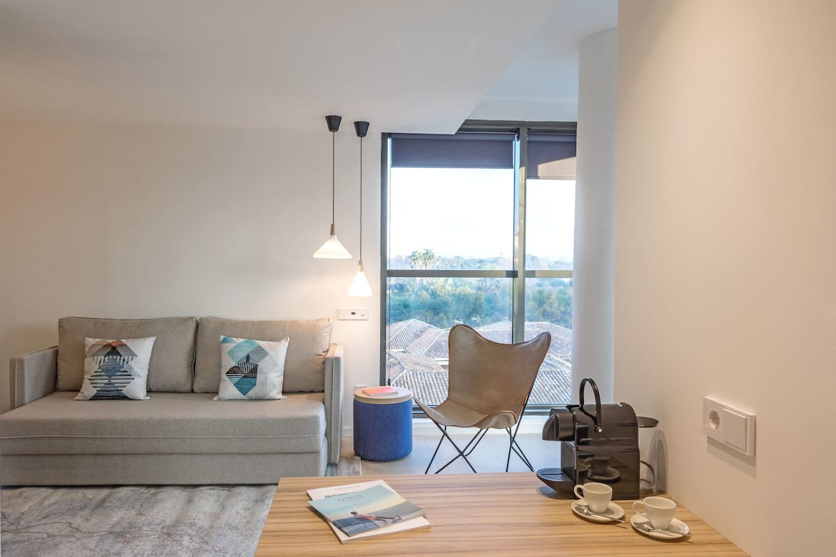 A modern living room with a sofa, a coffee table, a chair, and a view of a cityscape through large windows.