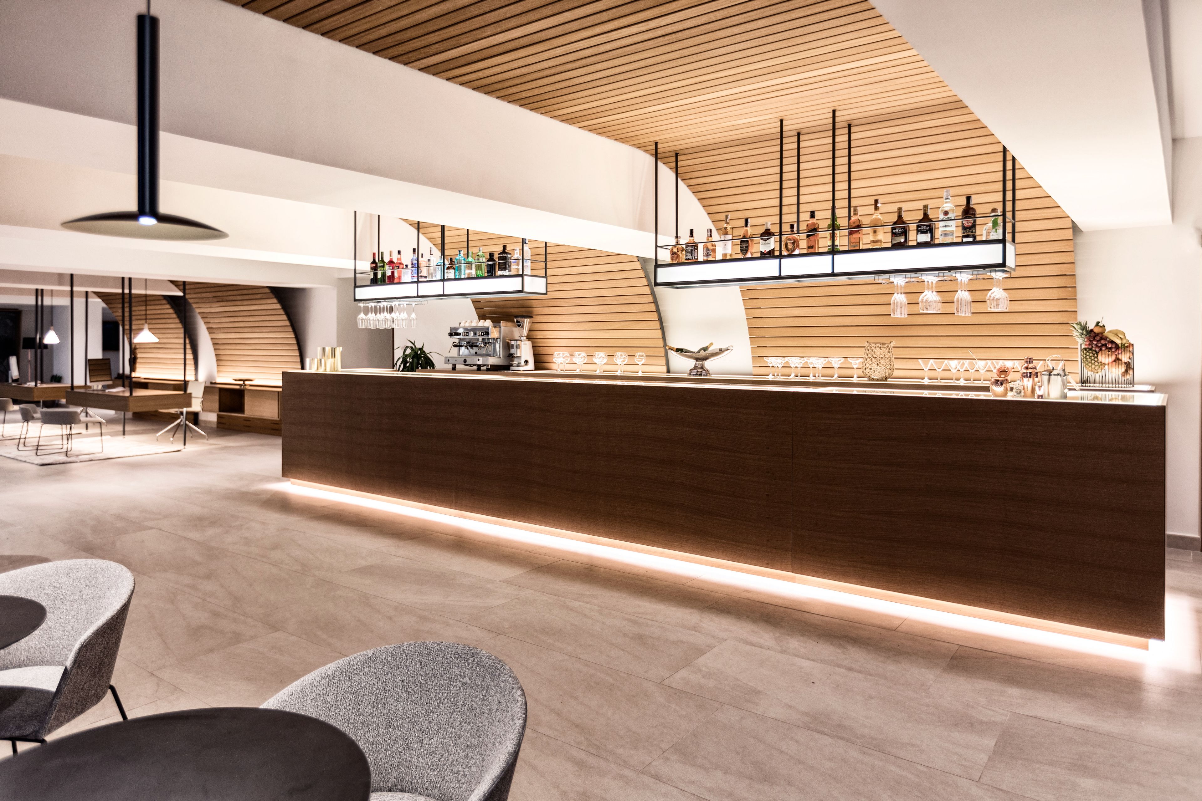 Zona de bar moderna con mostrador de madera, taburetes y una variedad de botellas y vasos.