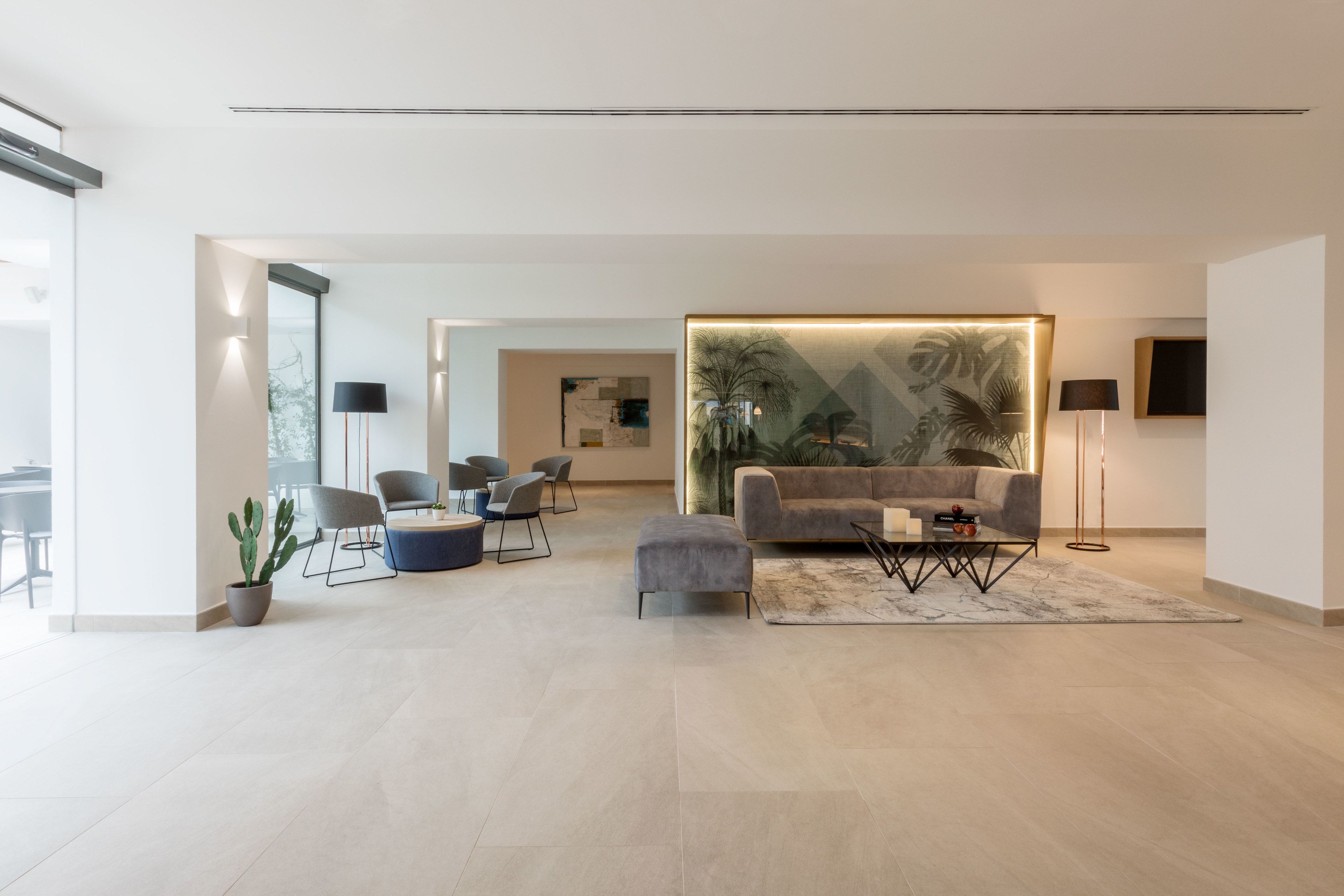 Una sala moderna con un sofá gris, una mesa de centro y una planta grande. Se ven varias sillas y una mesita en el fondo.