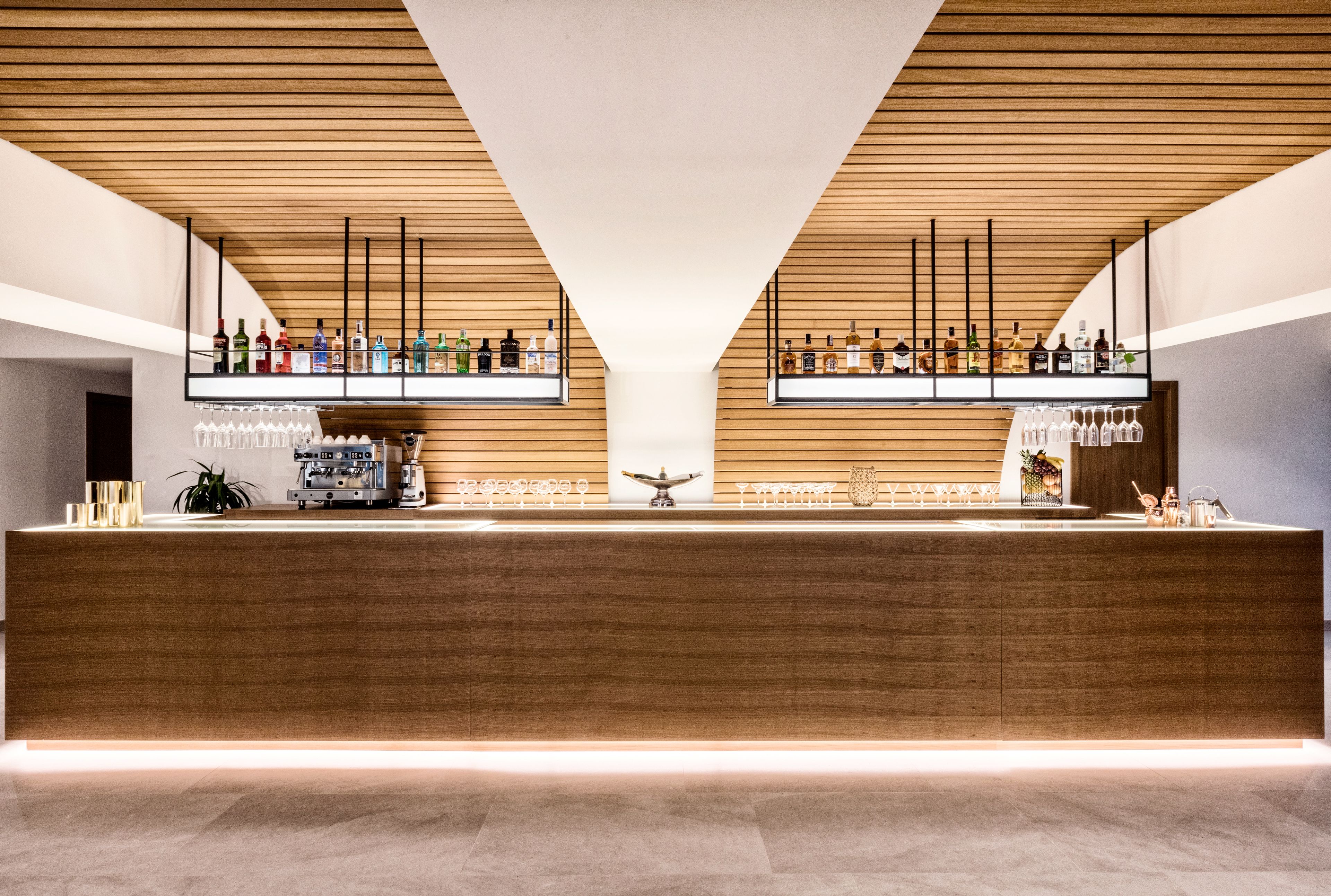 Un área de bar moderna con paneles de madera, una barra central y estantes con botellas y copas.