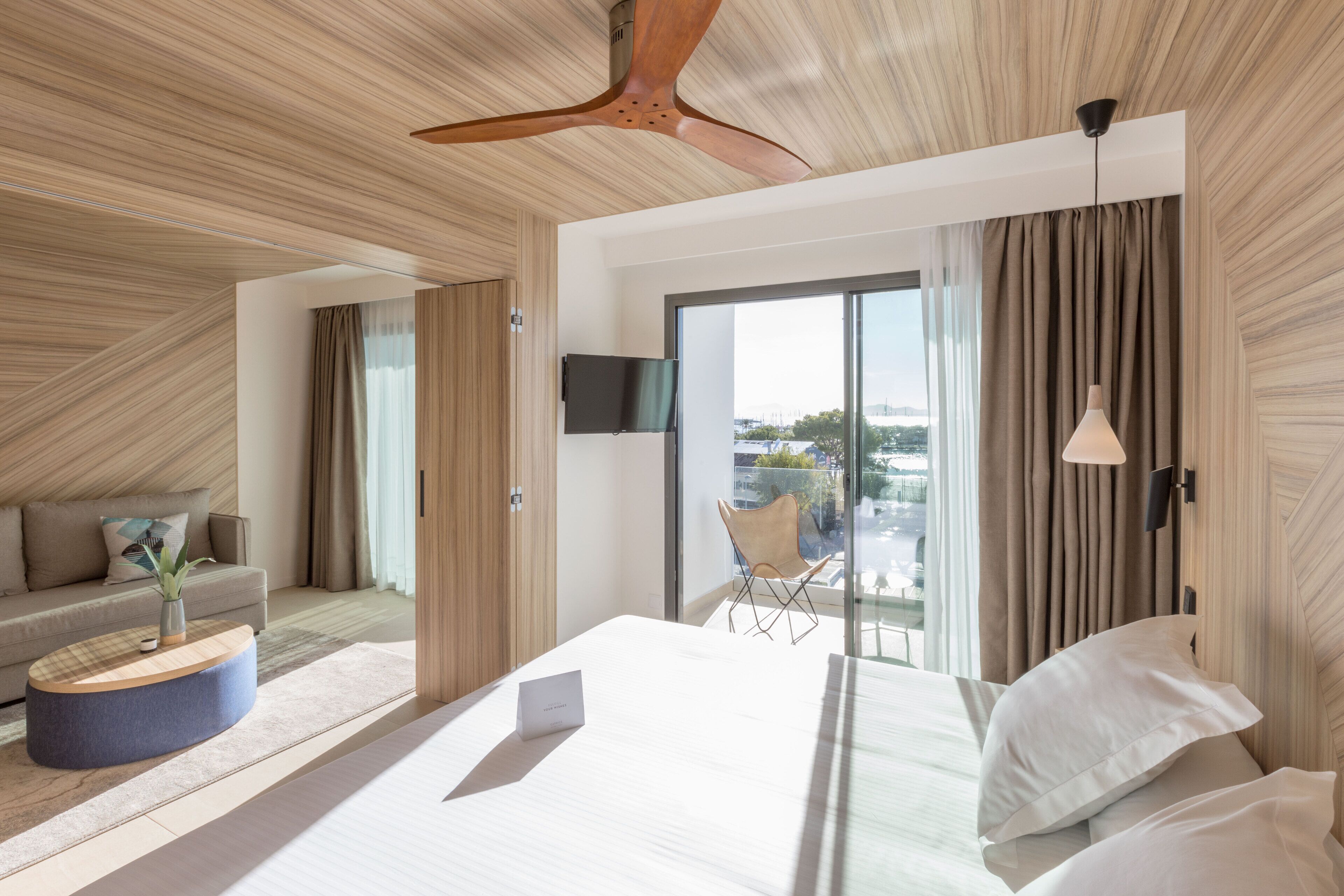 Una habitación de hotel moderna con una cama grande, un televisor de pantalla plana, un ventilador de techo y vistas a la ciudad a través de amplias ventanas.