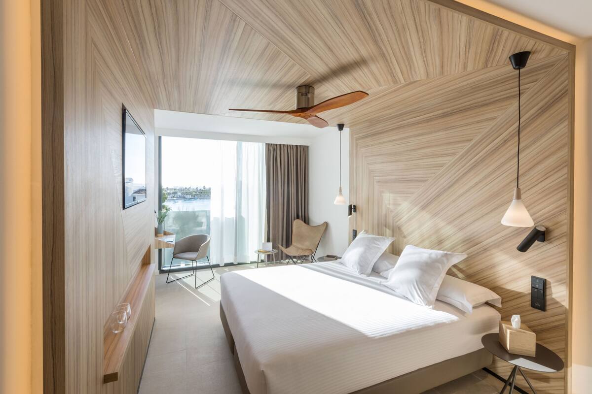 A modern hotel room with a large bed, a ceiling fan, a seating area with a chair, and a view of the city through large windows.