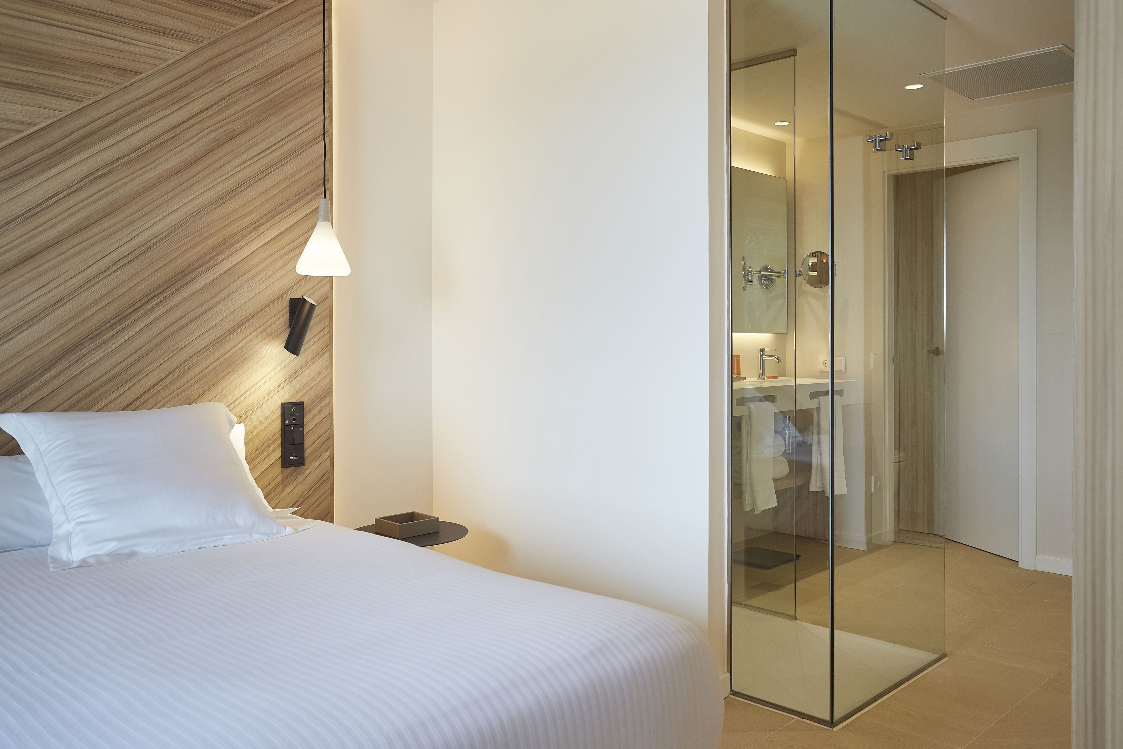 A modern hotel room with a large bed, a bedside table, a wall-mounted light, and a glass door leading to a bathroom.