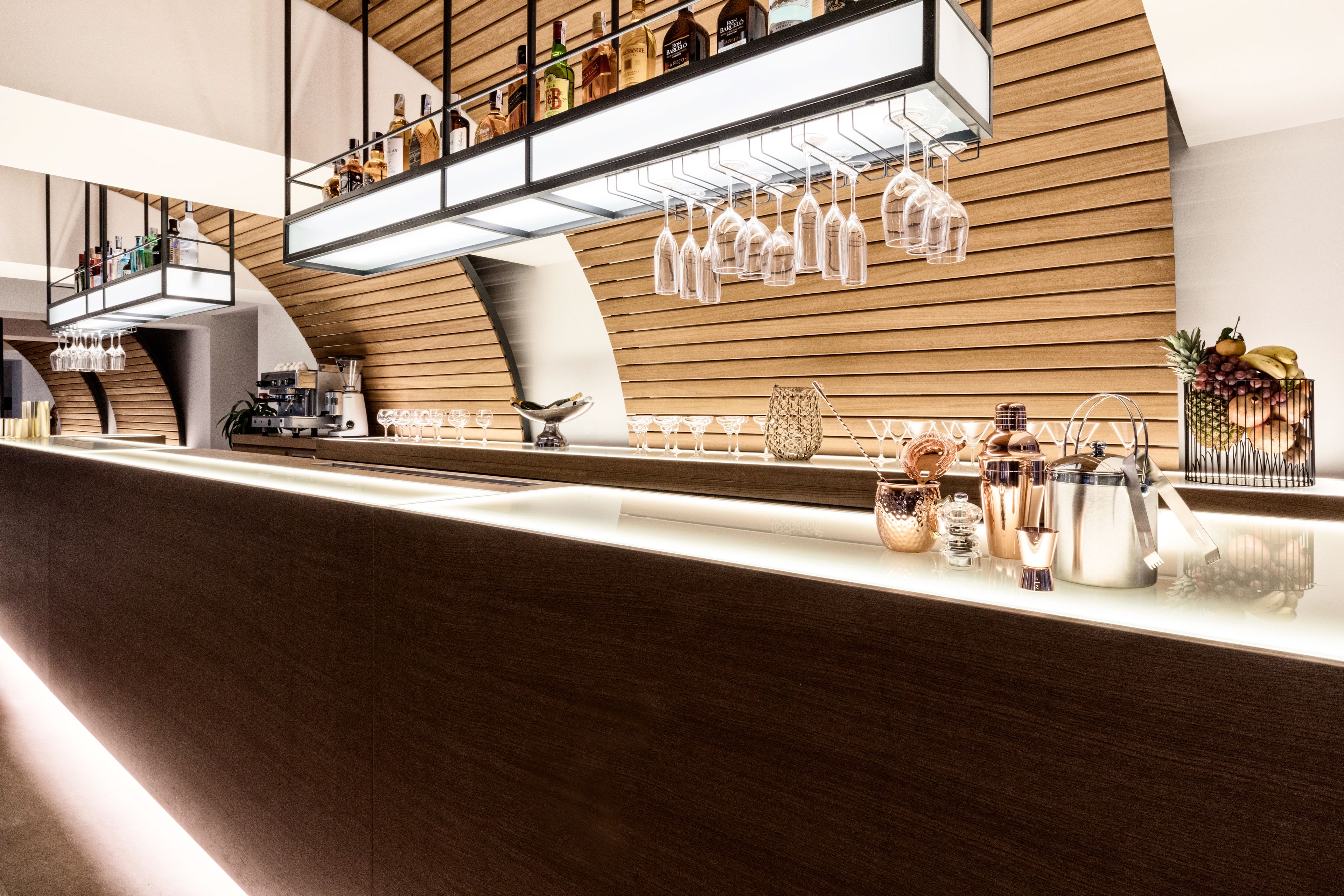 Zona de bar moderna con pared de madera, copas de vino, botellas y utensilios.