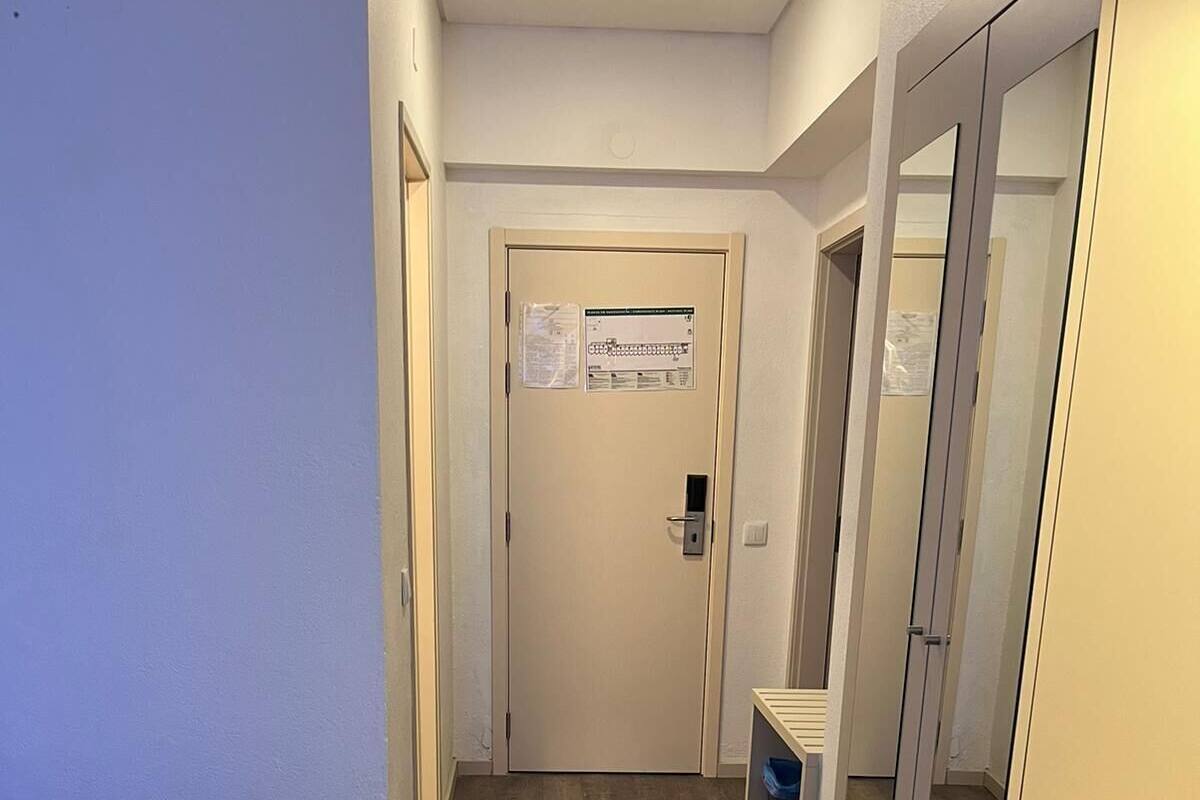 A hotel room with a bed, a desk, a chair, a trash bin, and a door with a sign.